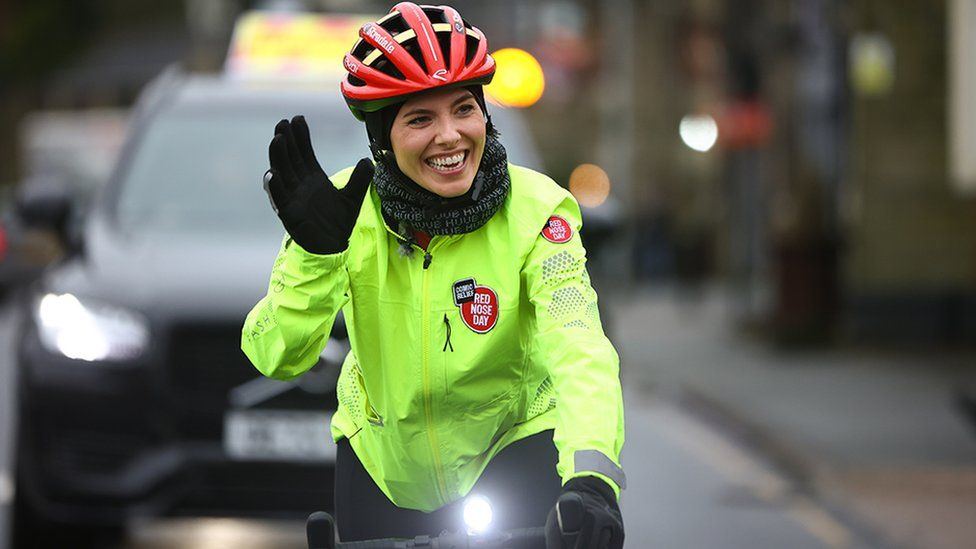 A woman rides a bicycle on a rain-covered road with an escort car trailling behind. She's smiling and waving, and wears a bright red crash helmet and a bright yellow hi-vis jacket. On the jacket a red circle with the words "Red Nose Day" written in white can be seen.