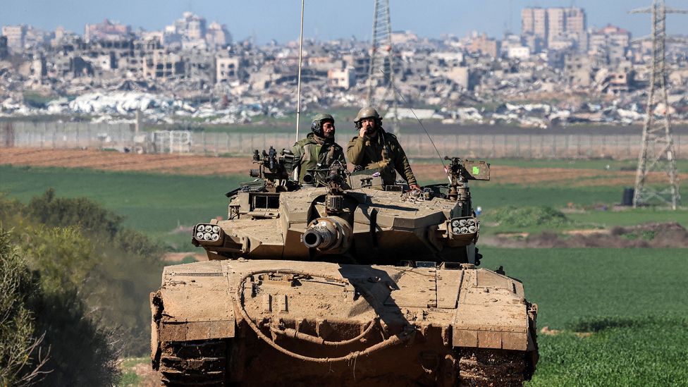 An Israeli army battle tank manoeuvres, with a landscape of damaged buildings in the background