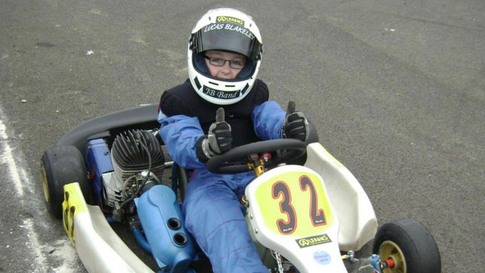 Lucas Blakely as a child, sits in a go-kart with the number 32 painted on the front. He's wearing a helmet but you can see his glasses, eyes and nose as his visor is up. He's wearing safety overalls and gloves, his hands are resting on the wheel in a double thumbs-up.