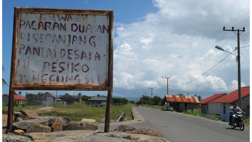 Водитель проезжает мимо таблички с надписью на местном языке: «Если вы встречаетесь на пляже, риск лежит на вас» в Банда-Ачех
