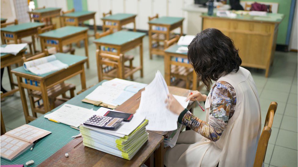A teacher sits at her desk marking in an empty classroom