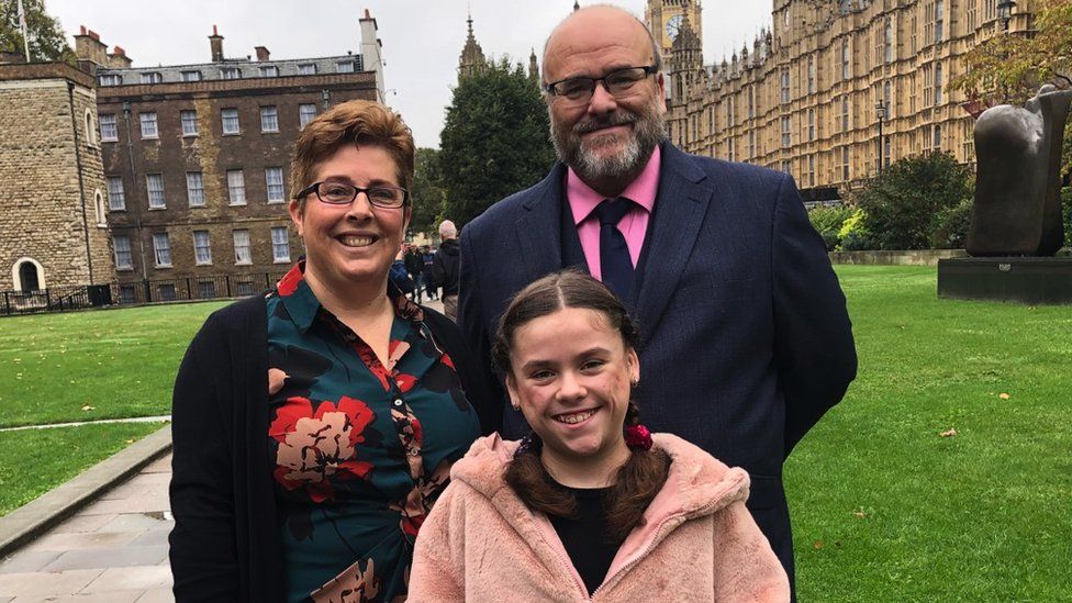 Carmela with her parents. Her mum is stood to the left of the frame. She has short brown hair and is wearing a green dress with red flowers. Her dad is on the right, dressed in a pink shirt and a navy blue suit and tie. He is wearing glasses and has a beard. Carmela is stood in front of them. All three are looking directly at the camera and smiling.