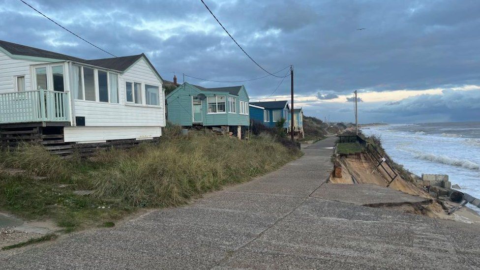 Wooden homes set back from concrete roadway which is being undermined by the sea.