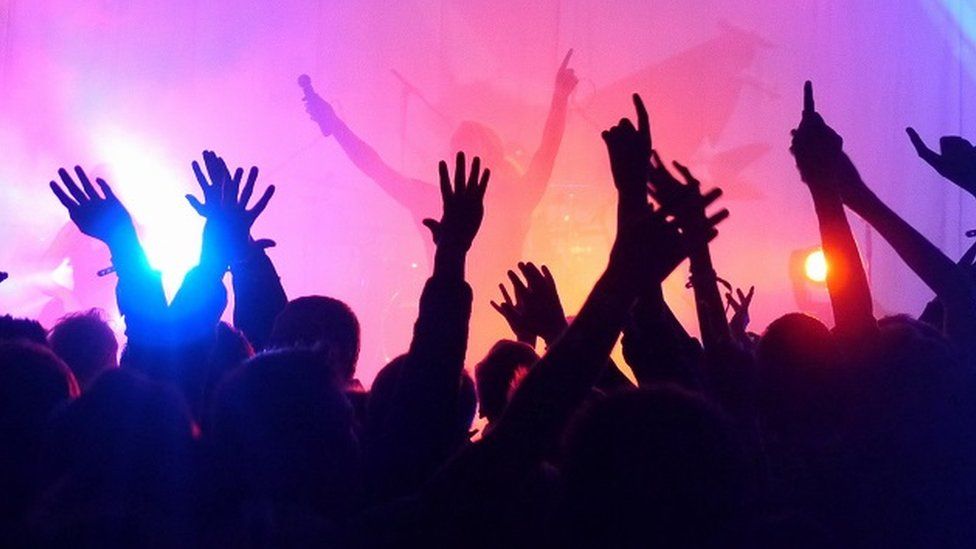 Stock image of people clubbing - there's a singer or dj on-stage with their arms raised, a microphone in one hand and the finger of the other pointing upwards. They're just visible through a sea of condensation or dry ice fog illuminated by pink and orange stage lights. In the foreground, silhouetted from the shoulders up, are 10 or 12 clubbers mimicking the DJ's pose.