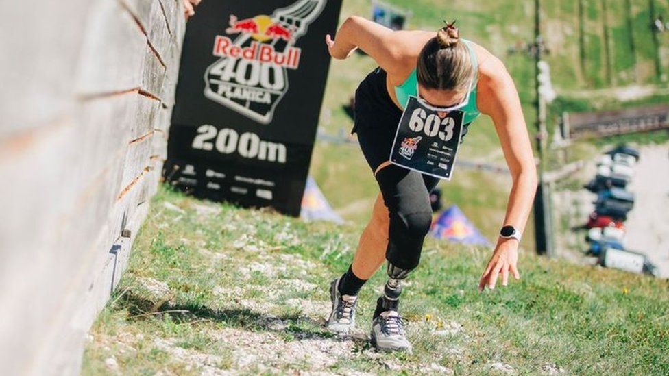 Milly Pickles at the 200m mark during the Red Bull 400 event in Slovenia. Millie, a white woman in her 20s, looks down as she strides up the incline. She has her brunette hair tied back in a bun and wears white framed sunglasses, a blue-green sports top and black shorts. She has a prosthetic limb on her right leg which is stretched in front of her left leg.