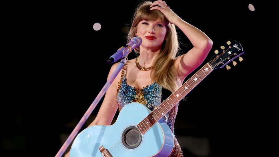 Taylor Swift on stage, she is wearing a blue leotard and has a blue guitar. Her hand is in her straight, blonde hair as she smiles