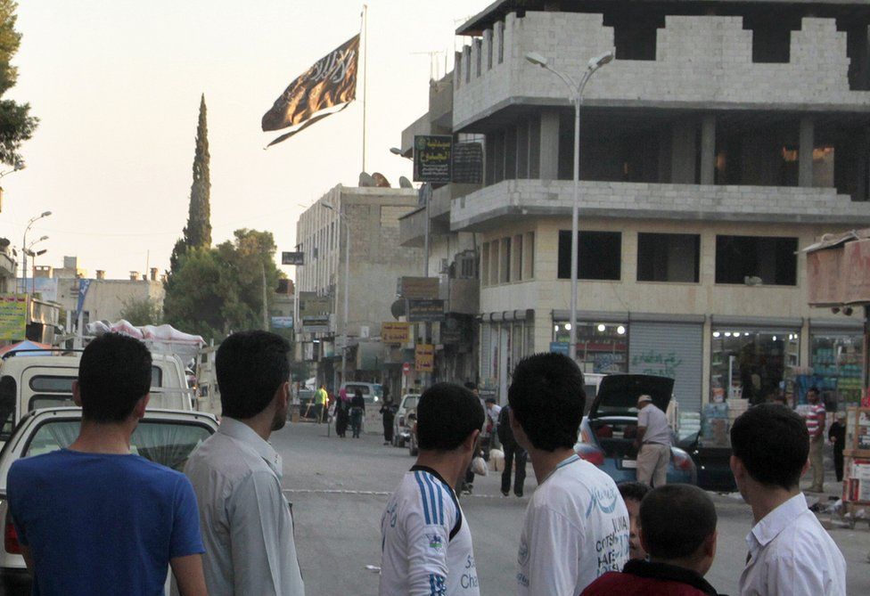 A group of men look at a large black jihadist flag with Islamic writing on it proclaiming, "There is no God but God, and Mohammed is the prophet of God".