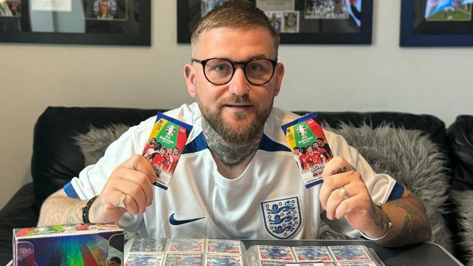 A man with large rimmed glasses kneels down over a table with a folder containing collectible football cards - each slotted neatly into a custom-designed protective pocket - opened out on it. He's wearing a white England football shirt and holding an unopened packet of Euro 2024 trading cards in each hand. On the wall behind him are three large frames containing signed football shirts and other memorabilia.