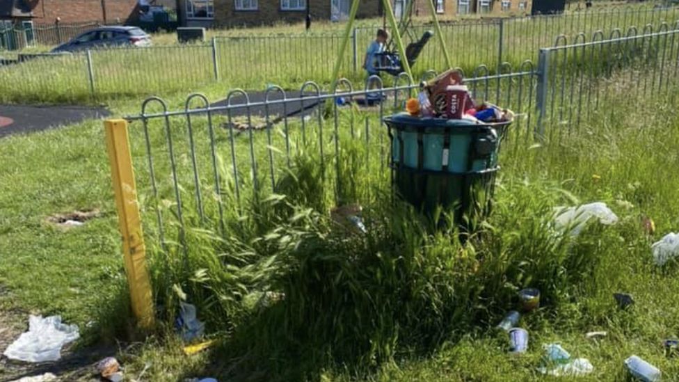 A bin is shown overflowing next to uncut grass by a playpark.