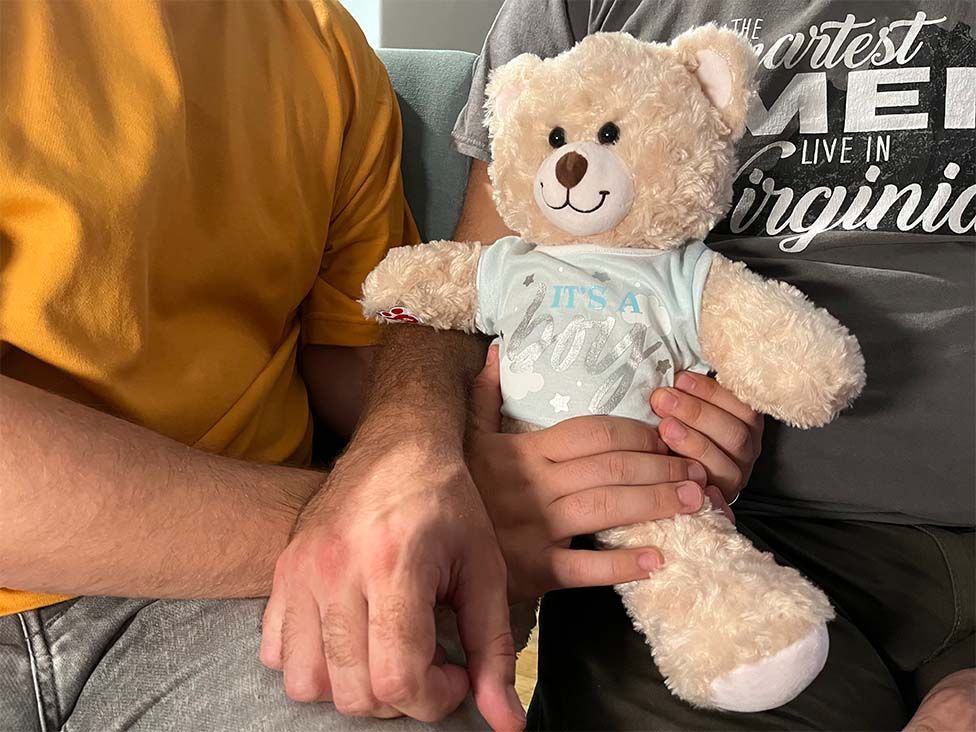 Two men holding a teddy bear wearing a top that says: It's a boy
