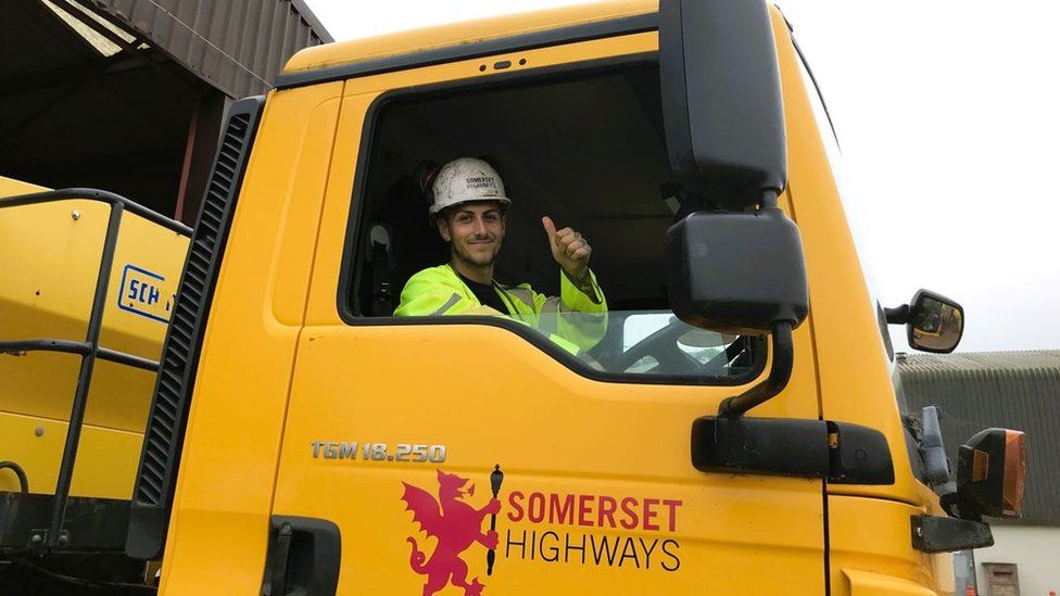 Josh From Somerset Council's Gritting Team wearing a hi-vis jacket and looking out the window of a gritter truck holding a thumbs up and smiling