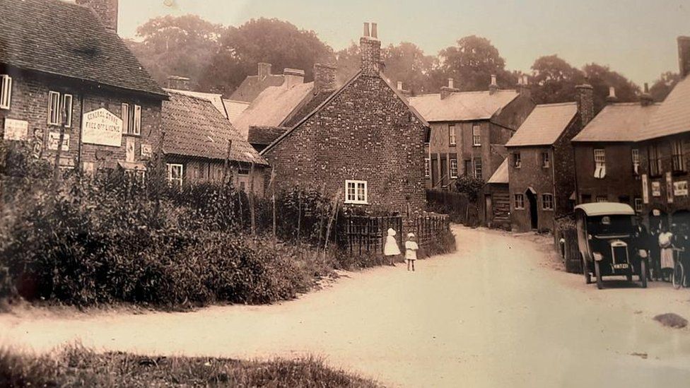 Black and white photo of village street showing shop and post office with old car outside