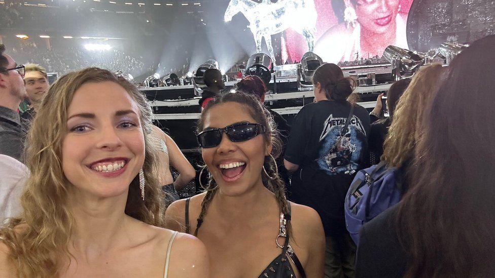 Alicia and Grecia standing in front of the stage, smiling. Grecia is wearing sunglasses and there are big bright lights pointing at the stage behind them,