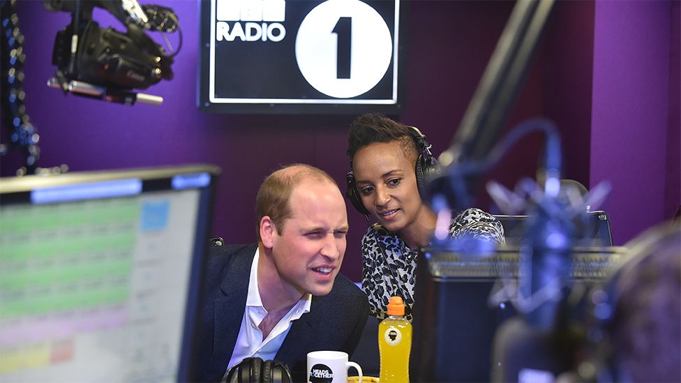 Prince William on the left and Adele Roberts on the right. The wall is purple, and the background has a sign reading BBC Radio 1, written in white and black background. A screen is on the let. There is a mug in front of Prince William, and a yellow plastic juice bottle in front of Adele. Prince William and Adele are both looking at another screen in front of them.
