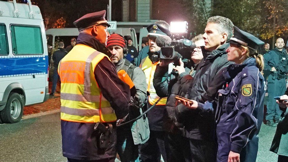 A police briefing at night is seen in this group shot, with one officer addressing a video camera which is professionally geared for audio
