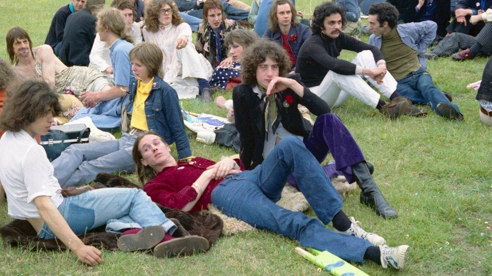 Peter Tatchell sits on the grass wearing purple flares and a black jacket