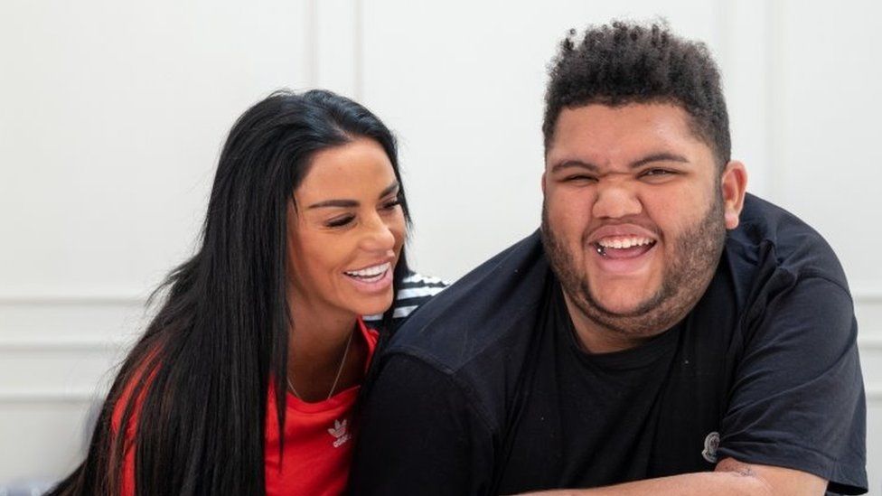 Image of Katie Price in a red top smiling while hugging her son Harvey, in a black t-shirt.
