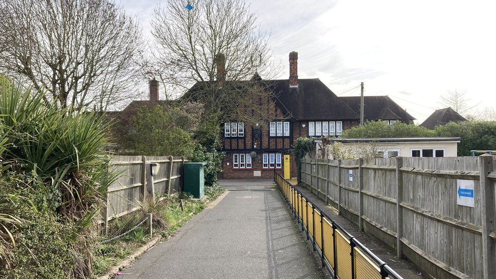 General view of the school, a mock Tudor building with a concrete pavement leading into a small fenced playground area