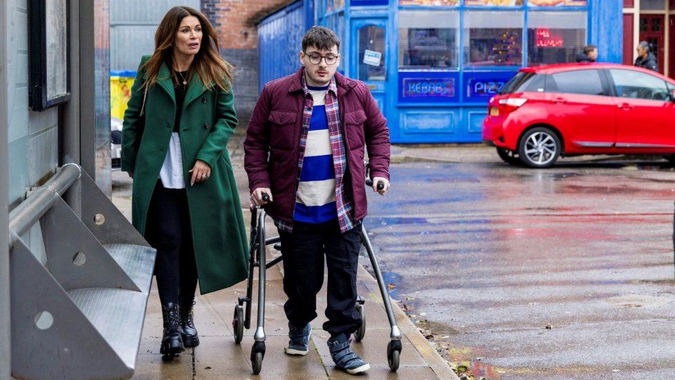 L-R Alison King as Carla Connor walks alongside Jack Carroll as Bobby on the Coronation Street set. Carla is a white woman in her 40s with long brown hair, she wears a long green coat over black trousers with black boots. Bobby, a white man in his 20s with short brown hair and dark rimmed glasses, wears a maroon jacket over a blue and white striped top with dark jeans and blue trainers. He uses a walker as the pair are pictured on a rain-soaked street in front of the set's kebab shop with a red car parked outside.