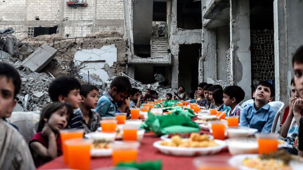 Children sit at a table as they wait for Iftar, evening meal at the end of daily Ramadan fast at sunset, next to rubble and destroyed houses, which were damaged after air strikes, in Douma, Syria, 17 June 2017.