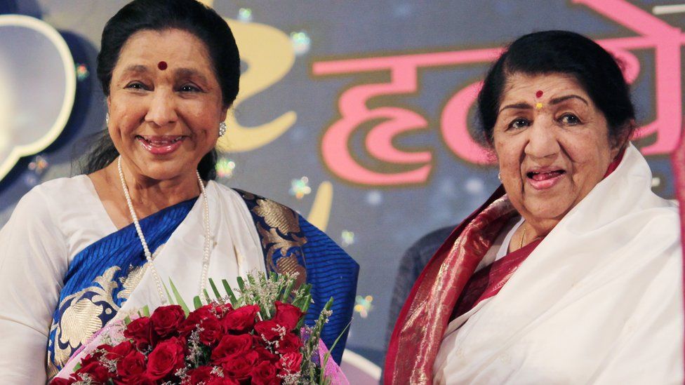 Asha Bhosle and Lata Mangeshkar holding flowers, two Indian women smiling, wearing white with red flowers in their hands. The background is pink writing in Hindi on a blue backdrop.