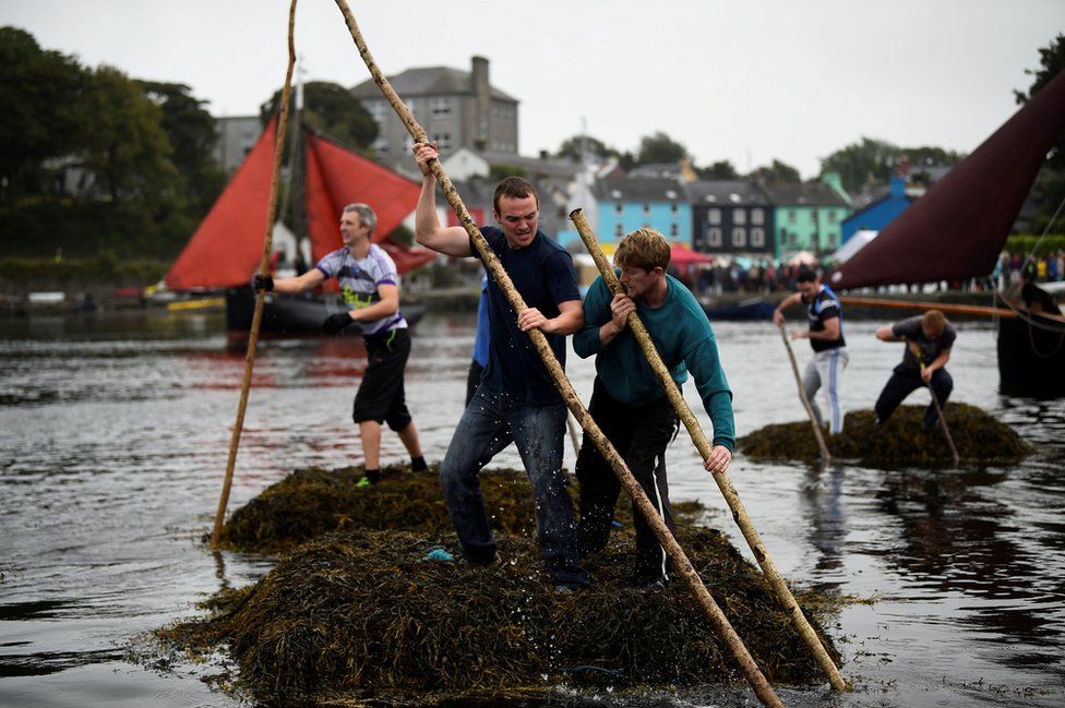 Two men push a seaweed raft using sticks.