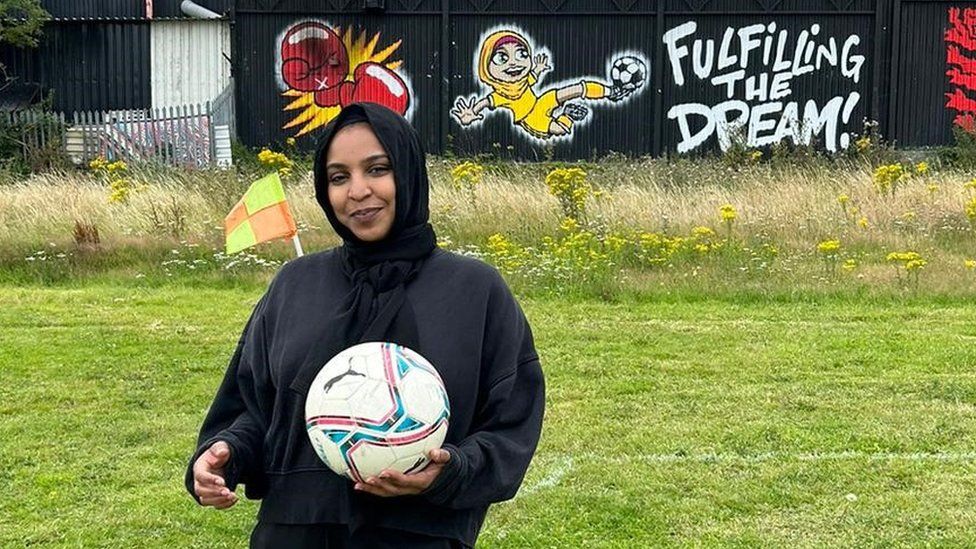 Asma Hassan standing in a grassy field holding a white football in her left hand. She is wearing a black hijab (headscarf) on her head and is smiling. She is wearing a black long-sleeved jumper. Behind her is a building with a mural on it that says "fulfilling the dream".