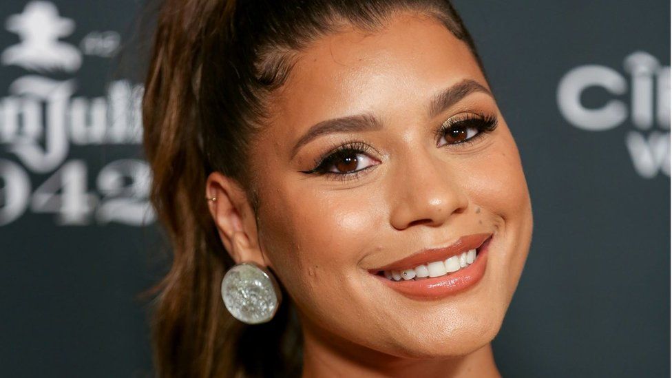 Milena Sanchez at an event in September. Milena is a black woman in her 30s, she wears her honey-brown hair tied back in a high pony tail. She smiles at the camera and wears large coin-shaped silver earrings.