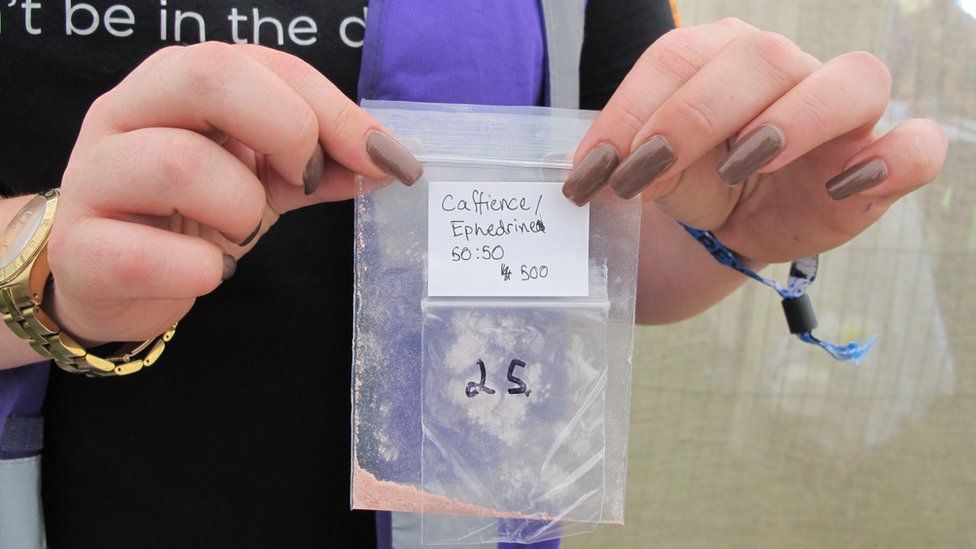 A close-up of a drug-tester's hands holding a resealable plastic bag with a pale orange powder at the bottom. A white sticker has the words "Caffeine/Ephedrine 50:50" handwritten on it. The drug tester has long fingernails painted chocolate brown and a festival wristband on one wrist