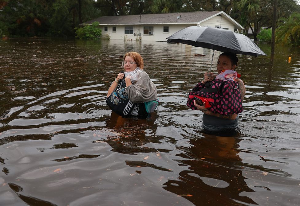Две женщины перебираются через паводковые воды после того, как им пришлось покинуть свой дом, когда паводковые воды от урагана Идалия затопили его 30 августа 2023 года в Тарпон-Спрингс, Флорида