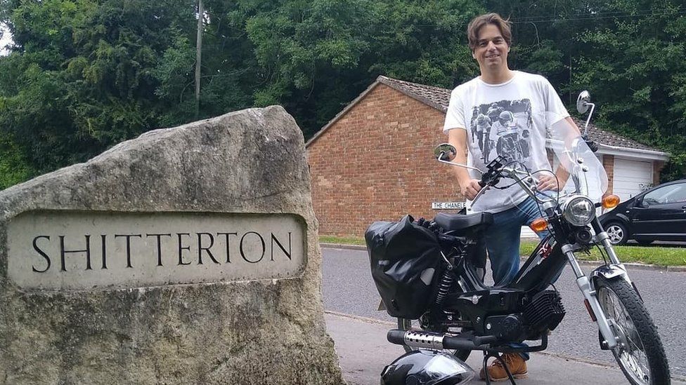 Paul Taylor with his moped next to the stone sign at Shitterton