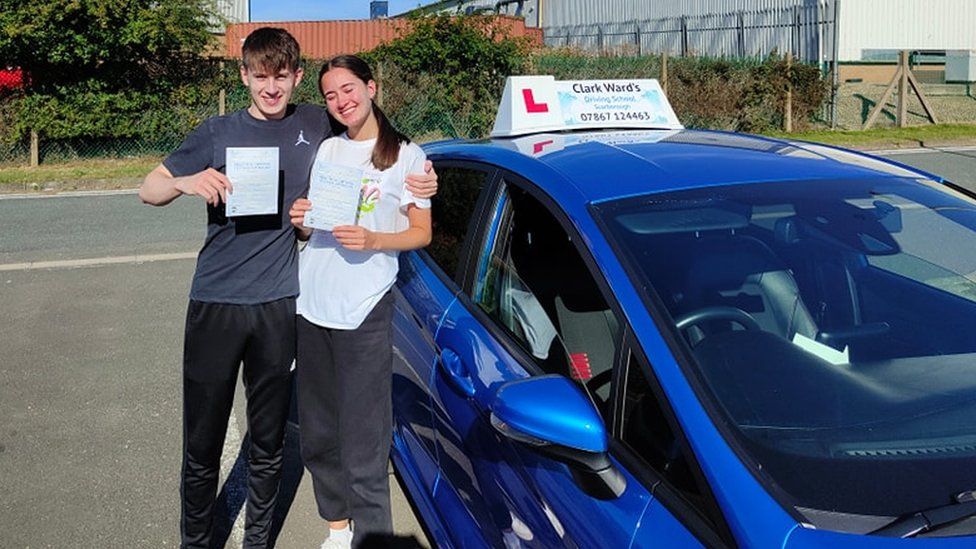 Twins holding driving certificates next to learner car