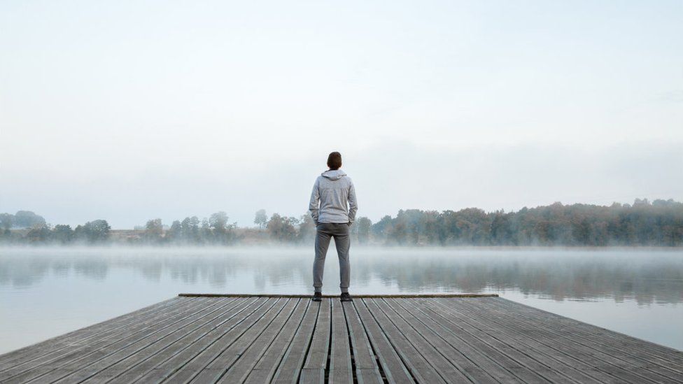 Young man standing alone on wooden footbridge and staring at lake. Thinking about life. Mist over water. Foggy air. Early chilly morning. Peaceful atmosphere in nature. Enjoying fresh air. Back view. - stock photo