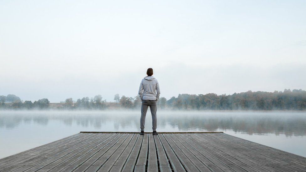 Joven de pie, solo, sobre una pasarela de madera y mirando el lago. Pensando en la vida. Niebla sobre el agua. Aire brumoso. Mañana fresca. Ambiente tranquilo en la naturaleza. Disfrutando del aire fresco. Vista trasera. - stock photo