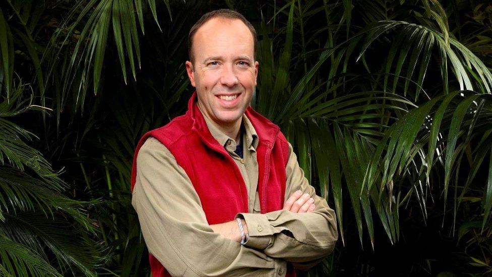 Matt Hancock dressed in beige shirt and red gilet stands arms crossed in front of jungle trees