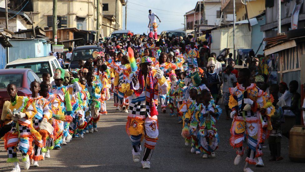 Lines of children parade in the street wearing multi-coloured feather outfits, led by an adult male. In the background, a man on stilts balances in the crowd in Sekondi Ghana