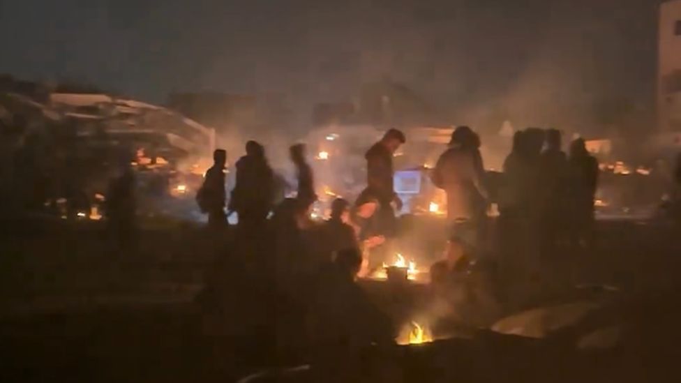 Still from an Instagram video showing figures waiting next to campfires in the dark.