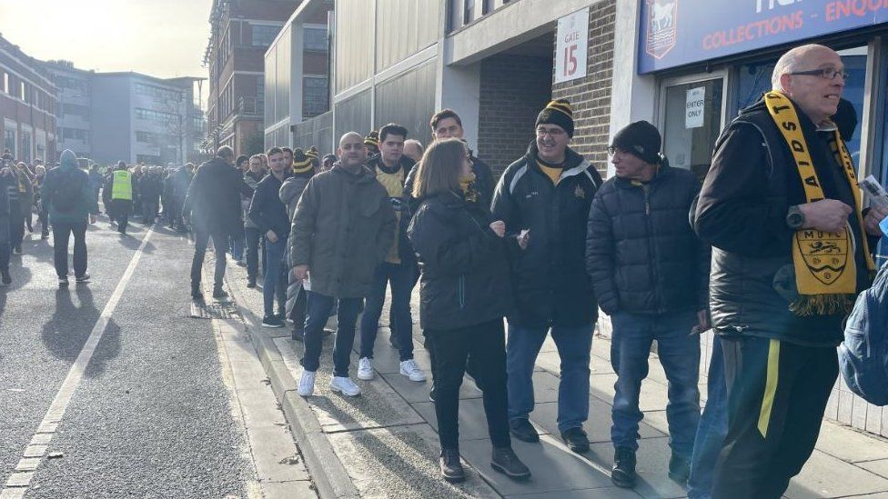 A queue of men and women wearing Maidstone United scarves outside the Ipswich Town Stadium
