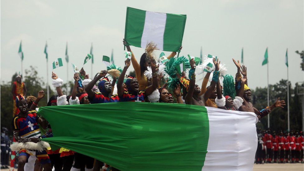 Members of an acrobatic band perform at Eagle Square in Abuja, Nigeria during the celebration for the country's 61st year of independence on Friday 1 October. There are a group of acrobats dressed elaborately with white face paint and wrapped in a large Nigerian flag jumping in a huddle. They look very happy and lively.