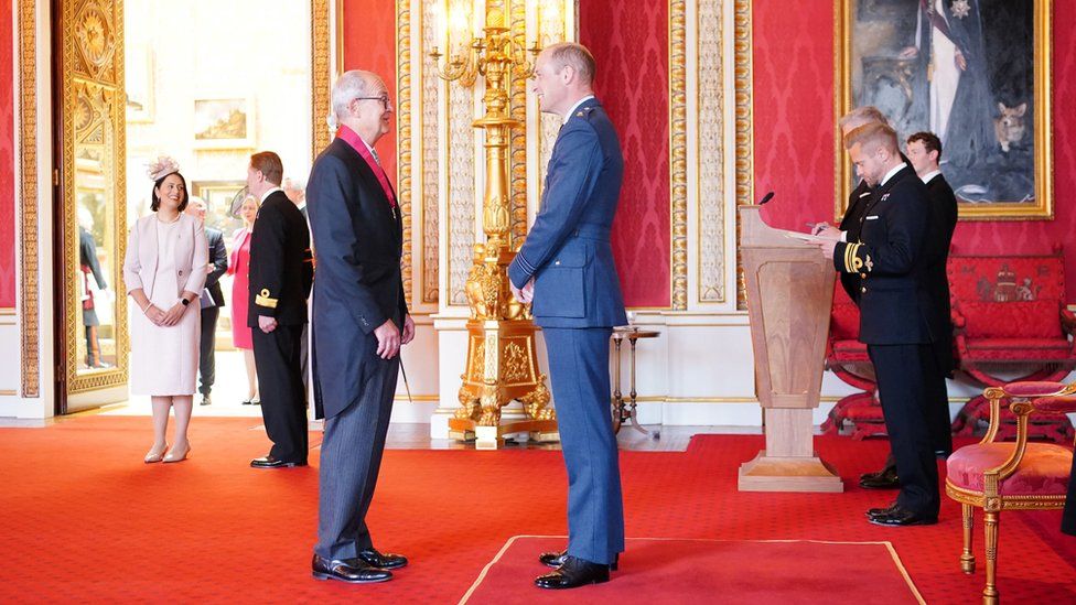 Sir Patrick speaking to Prince William as he receives his medal in an ornate red room in Buckingham Palace