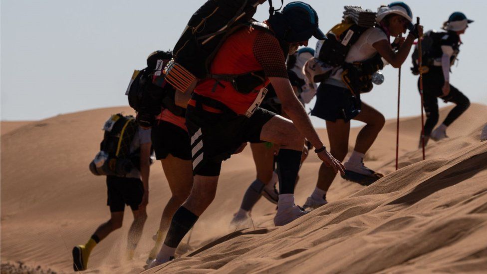 Marathon runners in running gear racing up a sand dune at incline. There are sand dunes in the background as well as relatively clear skies.