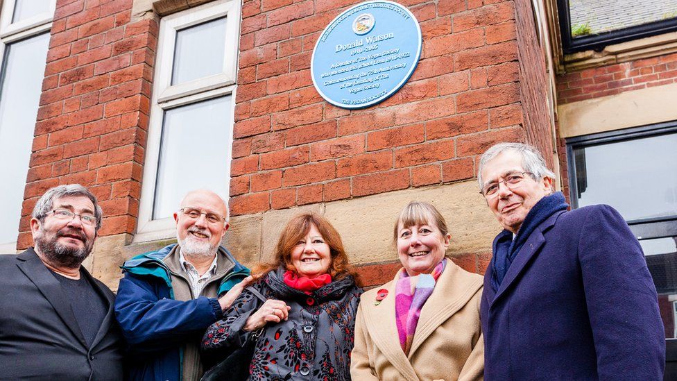 Five people standing below a blue plaque, which is on a building wall