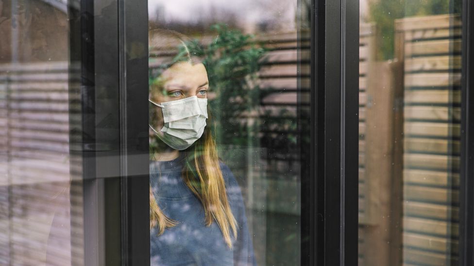 Woman stands behind window wearing face mask