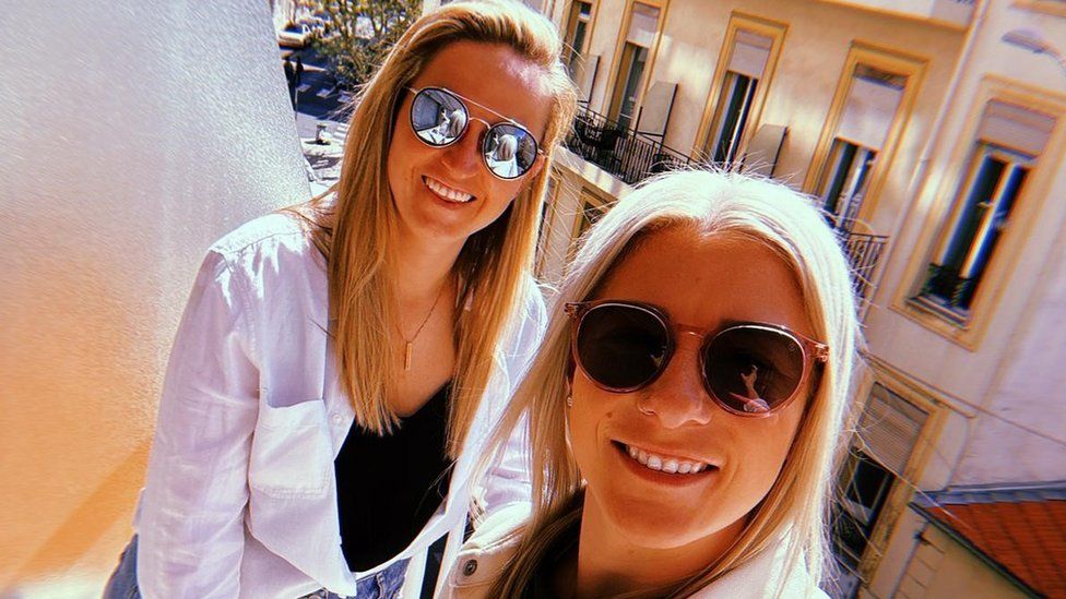 Bethany England with her fiancé Stephanie Williams. Bethany and Steph are both white women in their late 20s with long blonde hair worn loose - Bethany, on the right, wears hers centrally parted while Steph, left, has a side parting. They both wear sunglasses and are pictured smiling outside on a balcony in what appears to be a European city. Opposite other balconies can be seen across the street on a sunny day.