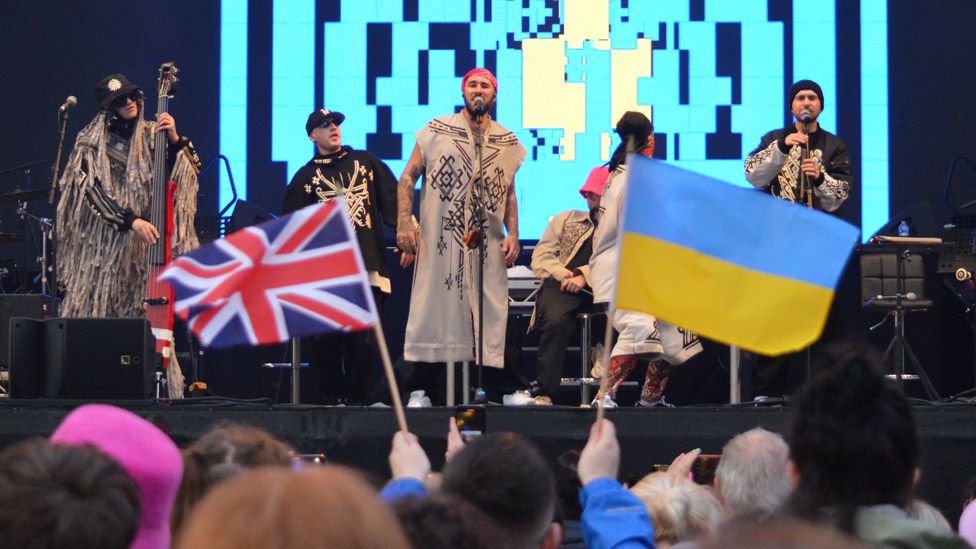 Kalish Orchestra on stage at the Eurovision Village in Liverpool, with UK and Ukraine flags in the crowd in the foreground