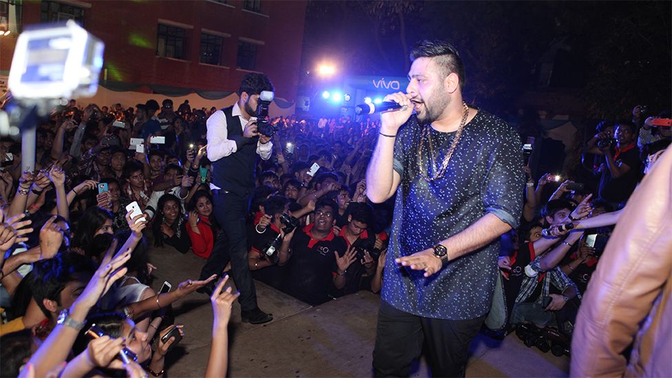Badshah giving a live concert performance at Delhi University. He is wearing a dark blie tshirt with small dotted patterns, and a silver chain around his neck. He is singing into a black microphone looking at the crowd, which are holding up their phones recording him. In the background there is a crowd in front of a bricked building, and a man wearing a white shirt, black waistcoat and trousers taking photos on a black professional camera. There are also blue flashing lights with a sign for the brand "VIVO" at the back.