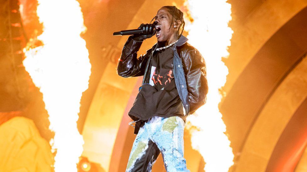 Travis Scott performs at Astroworld in 2021. He's wearing a black leather jacket, with a black hoodie underneath, and stonewash jeans. His gloved hand is holding a microphone close to his mouth as he roars into a microphone and jets of flame shoot out behind him on-stage.