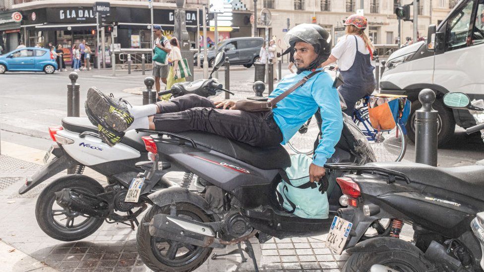 : Курьер еды отдыхает на своем мотоцикле на улице Канебьер, 12 августа 2023 года в Марселе, Франция. Марсель — второй по численности населения город Франции после Парижа