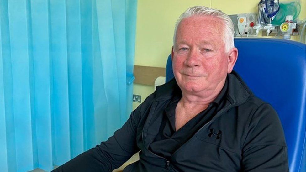 Scot Maclachlan sits in a hospital chair with a blue curtain next to him