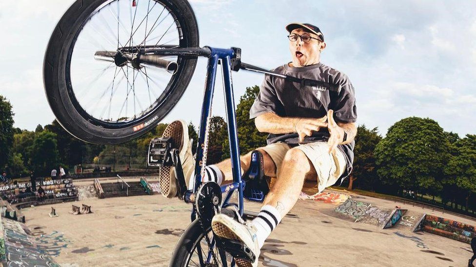 Dan Banks, a white man in his late 20s, pictured on his BMX bike mid jump at a skate park. Dan wears a black cap, round glasses, a baggy grey T-shirt and camel coloured knee length shorts, white trainers with white socks with two black stripes at the top. His feet are on the pedals however he's riding without his hands on the handle bars and pulls a shocked face as he looks at the camera. He rides a dark blue BMX bike with black wheels and behind him are trees, the skate park with ramps and graffiti and a blue sky.